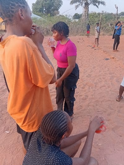 Three people are outside, possibly on a sandy area. One person wears an orange shirt and faces away from the camera. A person in a pink shirt drinks from a clear cup. Another person is to the right in a navy blue polka dot shirt, holding an orange item in their hands. Other people can be seen in the background, including someone wearing a blue shirt and black pants. There is a tree in the background.