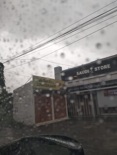 This media file captures a scene of a street in Banda Aceh, Indonesia, during a rainy day. The foreground shows part of a car, possibly a Suzuki based on visible text. The middle ground features two storefronts. The left storefront has a sign that reads "Oleh Oleh Khas SUMATERA BARAT," indicating it sells souvenirs from West Sumatra. This building appears to be a simple structure with closed brown doors. Adjacent to it, on the right, is a larger building with a prominent sign that says "SAUDI STORE" with a palm tree and sword logo. Below this, smaller text indicates offerings such as "Madu - Herbal - Oleh Oleh Timur Tengah - Perlengkapan ibadah" (Honey - Herbal - Middle Eastern Souvenirs - Religious Supplies). Another sign is partially visible that may say "Jamaah Yuk Ke" (Congregation, Let's Go). The weather is overcast, with visible raindrops on the lens, suggesting it is either raining or has recently rained. Power lines are visible strung across the sky, common in urban settings. The overall impression is of a typical commercial street scene in Indonesia, marked by the prevailing rainy weather.
