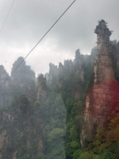 The image shows a landscape, possibly viewed through a window, featuring a mountain range with unique rock formations and lush vegetation. The sky is overcast, and there's a cable or wire running diagonally across the left side of the frame. The overall scene is somewhat obscured by mist or fog. The location is consistent with the Wulingyuan Scenic and Historic Interest Area in China, known for its sandstone pillars.