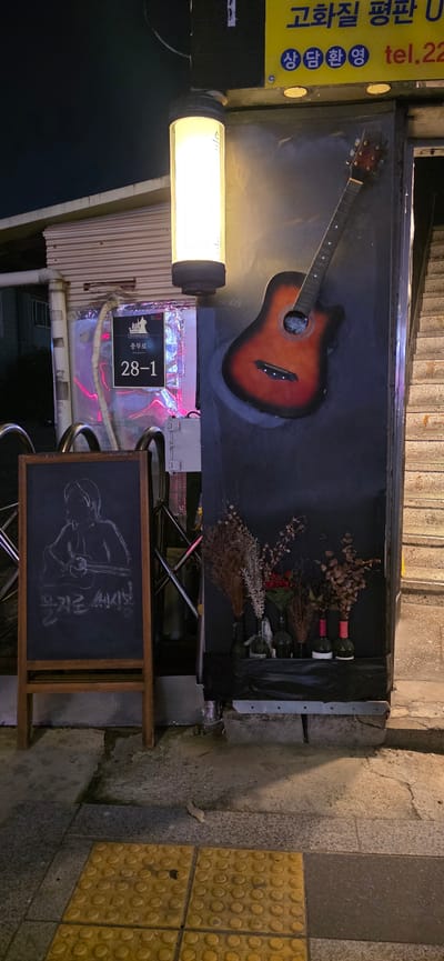This media file depicts a nighttime street scene in Seoul, South Korea, likely in front of a business. The primary objects in focus are a guitar mounted on a dark backdrop, several bottles of wine arranged with dried flowers, and a bright, cylindrical light fixture. A chalkboard sign with a chalk drawing of a person playing a guitar is also visible, leaning against a metal frame.
The sign to the left of the frame has Korean text and a phone number, possibly indicating a business offering services or products. Further details are visible on a sign attached to the building, which reads "충무로 28-1" (Chungmuro 28-1), providing a specific address.
The overall atmosphere is dimly lit, characteristic of a street at night. The presence of the guitar, wine, and chalkboard art suggests a possible music venue, bar, or a shop with a creative theme. The yellow tactile paving tiles on the right side of the image indicate a pedestrian area, typical of many city streets in South Korea. The image is captured at an angle, suggesting a casual, candid shot.