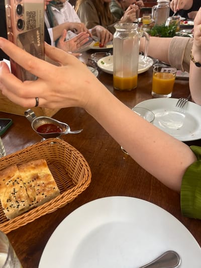 The image depicts a group of people seated at a wooden table, engaged in a meal. In the foreground, a person's arm is visible, holding a smartphone that has a payment card inserted into its case. A small metal dish containing a red sauce, likely ketchup or a similar condiment, is placed next to a woven basket holding two pieces of flatbread sprinkled with sesame and black seeds. Further back, other individuals are visible, with plates and glasses on the table. A glass pitcher filled with orange juice and a glass of the same beverage are also present. The background shows hints of other diners and their meals. The scene suggests a casual dining experience in a restaurant.