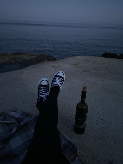 A person wearing black leggings and black and white Converse sneakers sits on a concrete surface next to the Adriatic Sea in Kunje, Montenegro. A partially consumed bottle of wine with a cork lies nearby. The person is resting on a plaid blanket. The environment suggests either dusk or dawn due to the dim lighting and low visibility over the water.