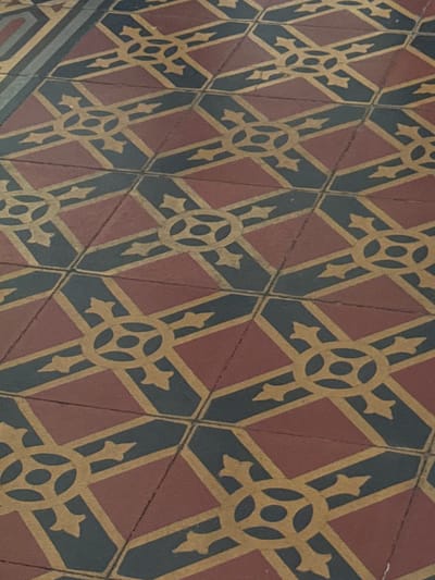 This image captures a close-up, oblique view of an intricate geometric tiled floor, likely located within a building in Catania, Italy. The floor features a repeating pattern composed of alternating reddish-brown and dark gray-blue (almost black) square tiles, laid in a diagonal grid.
Superimposed on these tiles is a detailed yellowish-gold pattern. This pattern consists of interconnected X-shapes, each centered around a circular motif that contains four smaller, symmetrically arranged circles or holes. Extending outwards from each central circle are four stylized, arrow-like or fleur-de-lis-like elements that form the arms of the 'X'. These X-shapes are linked by straight, diagonal lines of the same yellowish-gold, which also define the borders of the dark gray-blue tiles and traverse the reddish-brown ones.
The tiles show signs of age and wear, with visible grout lines and slight imperfections, suggesting the floor's historical nature. The lighting is even and diffused, not indicating a specific time of day. No people, objects, or activities are depicted, and there is no visible text. The overall design suggests a traditional or classical architectural aesthetic.