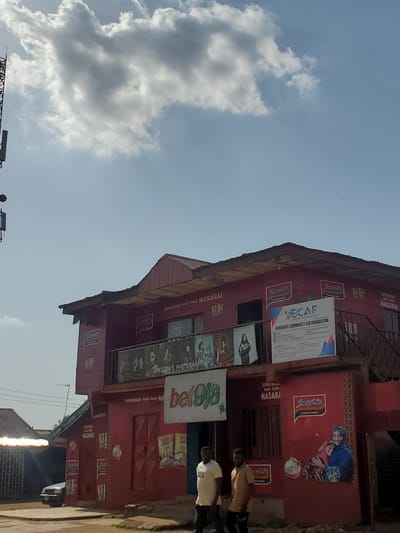 The image captures a street scene in Yelwa, Nigeria, featuring a two-story red building. The building is adorned with advertisements for "Indomie" instant noodles, "bet9ja," and the "BEMDOOSE COMMUNITY AID FOUNDATION (BCAF)." It also has signage for "PIXSORA'S PHOTOGRAHPY". The building is labeled with the phrase "Ingantaccen Abinci Domin NASARA!" on multiple locations. Two men are standing on the sidewalk in front of the building. The sky is partly cloudy.