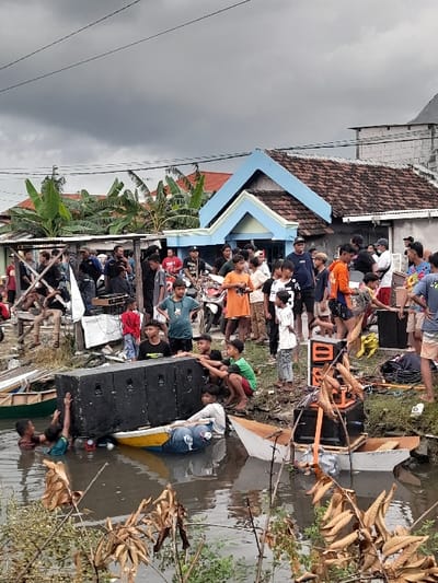 The image shows a scene of flooding in Kecamatan Tanggulangin, Indonesia, specifically at the coordinates -7.504 latitude and 112.746 longitude. There are numerous individuals, mostly men, standing, walking, and interacting in the flooded area. Some are carrying objects, others are looking at the floodwater, while some appear to be helping to move objects. There are also children in the scene. Some of the people are standing in front of a two-story house with a blue roof. Other objects in the scene include boats, large speakers, and other debris, such as chairs and pieces of wood, floating in the water. The water appears to be relatively shallow and brownish-grey in color. The sky is overcast with grey clouds. The environment is characterized by trees and foliage, suggesting a suburban or rural setting. The image conveys a sense of chaos and disruption caused by the flood.