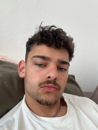 A close-up, angled portrait of a young man with dark, curly hair and a neatly trimmed beard and mustache. He is fair-skinned, has dark eyebrows, and appears to have greenish-gray eyes. He is wearing a white t-shirt and a silver chain necklace is visible at his neck. He is lying down on a soft, green surface (likely a couch or pillow). The background is a plain, white wall. There is no indication of the location being in Troyan, Bulgaria or any location.
