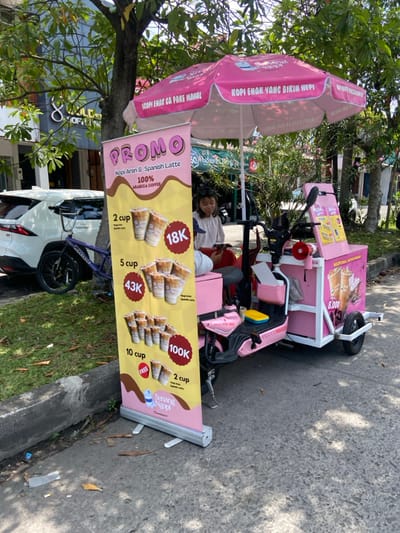 A pink motorized cart with an attached canopy, adorned with signs advertising "Kopi Enak" (delicious coffee) and "Promo Kopi Aren & Spanish Latte," is parked on a paved street in Margaasih, Indonesia (latitude: -6.963, longitude: 107.556). The cart displays a price list for various quantities of coffee, ranging from 2 cups for 18K to 10 cups for 100K. A woman in a blue shirt and black pants is seated on a small stool behind the cart, interacting with another person whose face is obscured. A bicycle is parked beside the cart, and a silver sedan is partially visible in the background. The cart is positioned beneath a large pink umbrella with the inscription "Kopi Enak Yang Bikin Heppy" (Delicious Coffee That Makes You Happy). The scene is bathed in sunlight, with a vibrant green tree providing shade in the background. The ground around the cart is strewn with leaves and debris, adding to the urban environment.