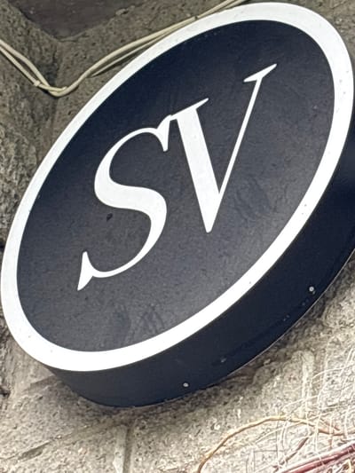 An angled view depicts a black oval sign with a white border, displaying a stylized, white "15". The sign is attached to a textured, light gray wall. A thin white cable is visible near the bottom left edge of the sign. The background includes portions of a textured wall and some dried vegetation. The location of Murino, Russia is not evident from this image.