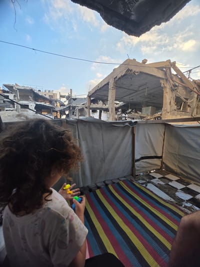 The image depicts a street scene in Gaza City, Palestinian Territories, characterized by widespread destruction. In the foreground, a young child with curly brown hair is seen from behind, wearing a light-colored t-shirt with a pattern of pastel-colored shapes. The child is holding several small, colorful plastic toys, including yellow and green pieces. The child is sitting on a striped mat, featuring alternating bands of red, yellow, black, and blue.

The middle ground shows a temporary shelter constructed from tarps and wooden supports. The tarps are light gray, with a black horizontal stripe on one section. The ground inside the shelter is partially covered by a black and white checkered tile pattern, with some areas appearing dirty or wet.

Dominating the background is a cityscape devastated by what appears to be bombing or shelling. Buildings are heavily damaged, with collapsed floors, exposed rebar, and gaping holes in their structures. Some structures show multiple stories of severe damage, with concrete debris scattered around. The sky above is blue with scattered white clouds. The overall impression is one of significant destruction juxtaposed with signs of everyday life, as evidenced by the child playing with toys.