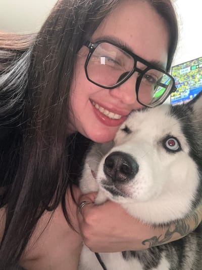 A woman with long, dark hair and glasses smiles as a husky nuzzles her face. The husky has striking blue and red eyes and black and white fur. A tattoo is visible on the woman's arm, which is being embraced by the dog. The scene appears to be indoors, with a television screen in the background displaying a sports broadcast, possibly a soccer game, with a score of "34:45" visible. The overall mood is affectionate and playful. The image is a close-up selfie, capturing a moment of bonding between the woman and her pet.