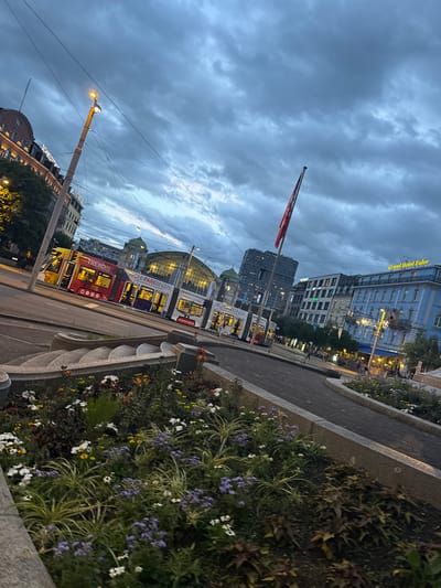 The image depicts a city street scene in Basel, Switzerland, likely near the Basel SBB train station. The sky is overcast with dark, dramatic clouds. A streetcar, decorated with advertisements for "TREND" and "WOHNPARKBINZEN," is visible in the foreground. Adjacent to the streetcar is a large building with a distinctive green dome, characteristic of the station's architecture. Several other buildings line the street, including a blue building with signage that reads "Grand Hotel Euler." A flagpole with a red flag stands prominently. In the immediate foreground, a flower bed with various colorful flowers and greenery is present. The overall atmosphere suggests dusk or a cloudy day, with artificial lights illuminating parts of the street and buildings.