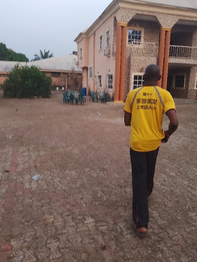 A man in a yellow shirt and dark pants walks away from the camera, toward a two-story building. The building has a peach facade with brown columns and a balcony. Green chairs and a blue container are grouped together in the building's yard, near a green bush and another building. The ground is a mix of brown dirt and sparse vegetation. The sky appears overcast.