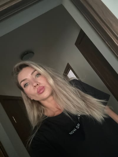 A selfie of a blonde woman is taken from a high angle, with her face positioned in the upper left quadrant of the frame. She is wearing a black t-shirt with white text that appears to read "NOW OR NEVER" in an arc. Her hair is long and blonde, and she is making a duck face. The background is dimly lit and appears to be an interior space with wooden trim and a ceiling light fixture. The setting is not identifiable as Armavir, Russia.