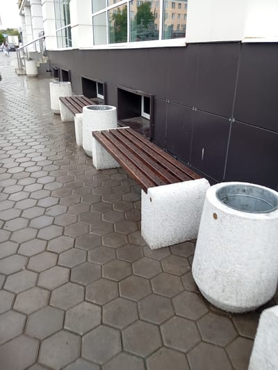 The image shows a row of outdoor seating arrangements along the exterior of a building. The seating consists of benches with wooden slats and concrete bases, and cylindrical concrete planters. The ground is paved with hexagonal grey pavers, which appear wet, suggesting recent rain. In the background, the building has a modern facade with dark grey panels and large windows. There are glimpses of buildings and trees visible through the windows. A metal railing runs along the left side of the scene. The image captures a public space, likely in a city. The presence of a white van with Russian license plates in the background, coupled with the architectural style and general urban landscape, is consistent with locations in the city of Orenburg, Russia. There are no people clearly visible in the immediate foreground or actively engaged in any interaction. The scene is static, depicting a street or walkway.