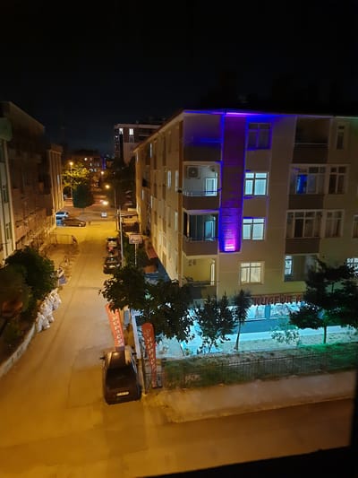 This image, taken at night, shows a residential street in the city of Atakum, Turkey. The street is lined with multi-story buildings, illuminated by streetlights and the lights from within the buildings. On the left side of the street, there are several white sandbags stacked against a building. further down the street, a dark-colored car is parked. On the right side of the street, a larger building is visible with decorative purple and blue lights along its facade. There is a storefront with text that appears to read "KUAFÖR A". Several trees and a fence line the area in front of this building. The overall scene depicts a quiet urban environment at night.