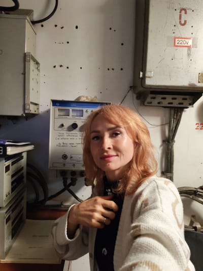 A medium shot shows a woman with shoulder-length auburn hair and fair skin posing for a selfie. She is wearing a cream-colored cardigan with a geometric pattern over a black top, and a ring on her left ring finger. Her right hand is touching her chest. The woman is positioned in front of a wall with several electrical panels and conduits. To her left, there is a white electrical cabinet with a black cable exiting the top. Next to it, a gray panel with a blue stripe is mounted on the wall, displaying two analog meters and various buttons and connectors. A handwritten note in Russian is affixed to this panel. To the woman's right, another gray electrical panel is mounted higher on the wall, with a red sticker indicating "220v" and a large red "C" above it. More conduits and wiring are visible on the right side of the image. The background suggests an industrial or technical environment.