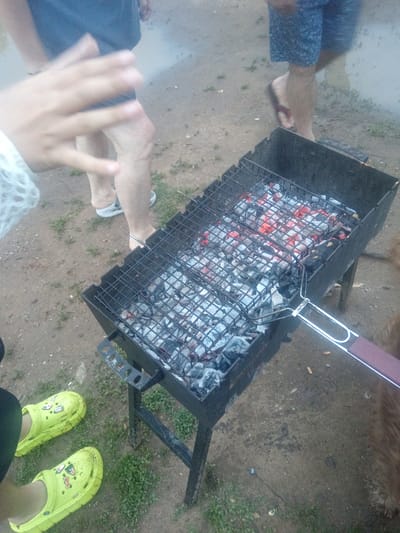 In Vasil'evka, Russia, a group of people are gathered around a charcoal grill. The grill is filled with hot coals, which are glowing red. A metal grate sits on top of the grill, ready for food. One person's hand is visible in the foreground, reaching out towards the grill. Their feet are clad in bright yellow Crocs. Another person stands with their back to the camera, wearing blue shorts. The ground around the grill is dirt and scattered with small green weeds. The scene appears to be an outdoor gathering, possibly a barbecue or cookout.