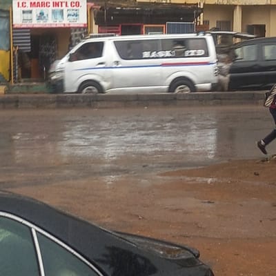 The image captures a street scene, likely during the daytime, with a wet ground surface suggesting recent rain or damp conditions. The setting appears to be a commercial or residential area.
In the mid-ground, a white passenger van, possibly a public transport minibus, is a prominent subject. It features distinctive blue and red stripes running horizontally along its side. On its side window, the word "BLACK" is visible in black text with a red outline, and "RED" is written in smaller red script above it, indicating a potential route or company name ("BLACK RED"). The van's wheels are visible, and it appears to be parked or moving slowly.
To the far right, the front passenger side of a black car, including part of its window and roofline, is partially visible.
A person is seen in the upper center of the image, walking away from the camera. They are wearing dark trousers and dark shoes.
On the left side of the street, a building is visible, and near its entrance, several rectangular objects resembling solar panels are displayed. Below these, a white sign stands vertically, bearing the text "I.C. MARC INT'L CO." in red letters. Smaller, less discernible text and images are also present on the sign. The overall scene depicts typical daily activity on a street.