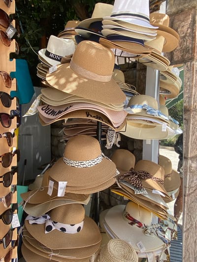 In Petrovac centar, Montenegro, a stall displays a variety of straw hats and sunglasses. The hats are stacked in multiple tiers, with the majority being wide-brimmed and made of woven straw. Several hats are adorned with ribbons in various patterns, including polka dots, abstract designs, and floral prints. Some hats also feature lettering, such as "MONTE" on a white fedora.
To the left of the hats, a wooden rack holds several pairs of sunglasses. These sunglasses have dark lenses and frames in shades of brown and black. The background of the scene consists of a stone wall, lush green foliage, and hints of a building's exterior. Sunlight casts shadows on the hats, indicating an outdoor setting. The overall impression is a retail display, likely in a tourist-oriented area.