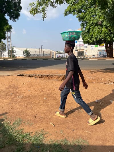 In Unguwar Sarki, Nigeria, a young boy walks along a dirt path beside a paved road. He is carrying a green plastic basket filled with items on his head. He is wearing a black t-shirt with pink accents on the sleeves and dark denim jeans that are ripped at the knees. His feet are shod in light yellow slides. The ground is a reddish-brown dirt, with patches of green grass growing along the edge of the path. In the background, across the road, is a large white building with a fence and a gate. A row of trees with green foliage flanks the building. The sky is blue with scattered white clouds.