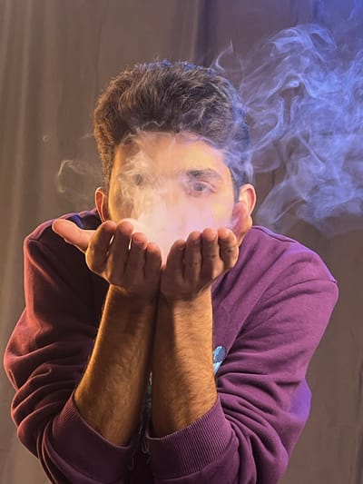 A person in a purple sweatshirt holds their hands up to their face, exhaling a cloud of white and blue smoke. The smoke obscures the lower portion of their face, while their eyes are visible above. The individual has short, dark hair. The image is angled, giving a slightly disorienting perspective. The background is a neutral, blurry gray.