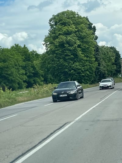 The image shows a road scene in Osenets, Bulgaria. Two vehicles are present: a black car closer to the viewer and a white vehicle further ahead. Both cars are traveling on the road. Lush green trees line the side of the road, and a partly cloudy sky is overhead. The road is asphalt with painted white lines.