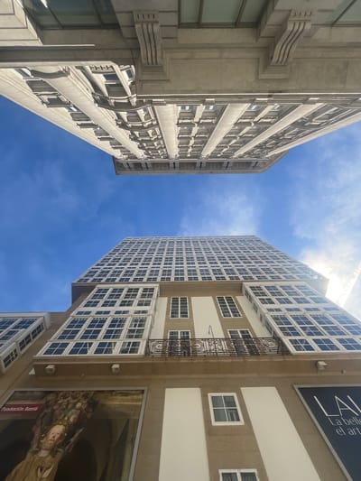 The image shows a low-angle shot of the Maria Pita Square in A Coruña, Spain, featuring the imposing architecture of the City Hall building. The square is famous for its iconic circular design and the statue of María Pita at its center, although these are not directly visible in this particular frame. Instead, the focus is on the facades of the surrounding buildings, which are characterized by their traditional Galician style with large glassed-in balconies, known as "galerías." The sky above is a vibrant blue with scattered white clouds. One of the buildings displays a large mural art piece on its lower level, and a sign partially visible reads "Fundación Barrie." The architecture is grand and ornate, reflecting the historical significance of the location.