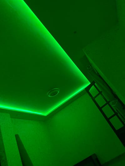 The image depicts an interior view of a room, predominantly illuminated by green LED lighting. The lighting is integrated into a recessed ceiling, creating a luminous line along the edges of the ceiling. A circular light fixture is visible on the ceiling. The walls have a textured finish. In the upper right portion of the image, a doorway with a dark-colored frame and glass panels is visible. The scene is devoid of any individuals or specific activities. The location is identified as Baraki, Algeria.