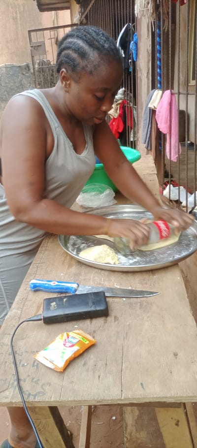 An adult female is observed preparing food outdoors in Ogidi, Nigeria. The subject, with dark skin and braided hair, wears a grey tank top. She is actively pressing and rolling a yellowish dough on a large, round, metallic tray, utilizing an empty Coca-Cola plastic bottle as a rolling pin. On the wooden surface in front of her are a knife with a blue and white handle (featuring a red star emblem) and a silver blade, a black power bank with an attached USB cable, and an orange and white sachet. The background features a dark metallic gate draped with various pieces of clothing in multiple colors (red, blue, pink, white), a green plastic basin, and a clear plastic bag. An earthy brown wall is visible behind the gate, with a corrugated roof section in the upper left. The ground is reddish-brown earth.