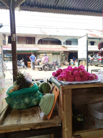The image depicts a street scene in Mns Alue, Indonesia, viewed from under a corrugated metal awning. In the immediate foreground, a wooden market stall displays produce: a green mesh bag contains several light-colored, round fruits (possibly coconuts or melons) with leafy stalks, alongside two large watermelons. To the right, a white tray holds a substantial pile of individual fruits, each encased in a pink protective foam net. A light green colander and an orange cutting board rest on the wooden surface. A wooden post with a white electrical outlet is visible on the far left.
The midground features a paved street with multiple individuals and vehicles. Three men are central: one in a blue shirt and dark pants stands upright, another in a light-colored shirt bends over a low object or cart, and a third in a purple shirt stands near a parked black motorcycle. Another lighter-colored scooter is parked to the left. Further down the street on the right, a black car is partially visible. Multiple small street-level shops or stalls, identifiable by their corrugated metal awnings and various goods, line the street. A white signboard with "BON KRISTA" text is visible on the right, near an arrangement of dark, hanging produce.
In the background, two-story buildings with predominantly wooden facades, arched openings on the upper levels, and corrugated metal roofs line the opposite side of the street, consistent with local architecture. The scene is illuminated by bright daylight, indicating an outdoor, active market environment.