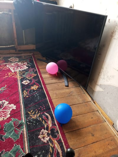 A large, black, flat-screen television is tilted on its stand against a wall in what appears to be a room with wooden floors and a patterned rug. Two balloons, one pink and one blue, are on the floor near the television. The wall behind the television is textured and appears to be wallpapered. The overall scene suggests a casual, indoor environment. The location is Vsevolozhsk, Russia.