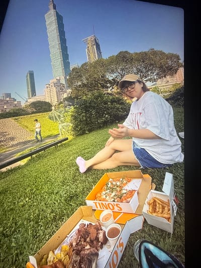 The image depicts a casual outdoor scene with a woman sitting on a grassy lawn, holding pizza boxes and french fries. In the background, the iconic Taipei 101 skyscraper and other city buildings are visible under a clear blue sky. The woman is wearing a white t-shirt, denim shorts, a beige baseball cap, and glasses. Two open pizza boxes and a container of french fries are placed in front of her. A path and trees are also present in the background, with a person walking in the distance. The overall setting appears to be a park in Taipei, Taiwan, offering a view of the city skyline.
