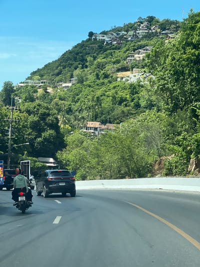 This image depicts a road scene in Baan Chaweng Noi, Thailand. In the foreground, a dark-colored car and a motorcycle are traveling on a paved road with a yellow dividing line. A person wearing a light green jacket and an orange helmet is riding the motorcycle, which has a license plate with Thai characters and numbers. The car has a license plate that reads "5BD 6204". In the background, a lush green hillside is covered with dense vegetation and numerous houses, many of which appear to be villas or resort accommodations, built at various levels up the slope. The sky is clear and blue. The environment suggests a tropical, hilly coastal area.