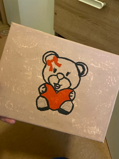 A close-up image depicts a rectangular object, likely a canvas or box lid, held by a human hand in an indoor setting. The object's surface is a speckled, light beige hue. Dominating the center is a hand-drawn illustration of a teddy bear, outlined in black, with a light beige body. The bear features a red bow on its head and holds a solid red heart in its paws. Its face includes two small black eyes, a black nose, and a smiling mouth with a pink inner detail. The paws display black pads with four lighter outlines suggesting toes. A partial human hand, showing a thumb and index finger, grips the lower-left edge of the object. The background is out of focus, revealing a light brown wooden surface with a white horizontal drawer pull in the upper right. Other blurred areas suggest indistinct interior elements. The scene is located in the city of Senden, Germany.