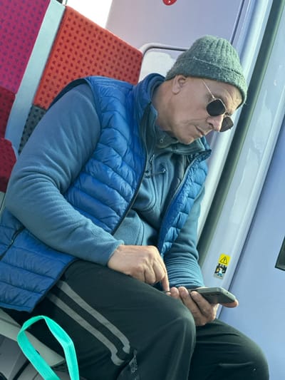 An adult male is depicted seated within a public transport vehicle, consistent with a train or subway carriage operating in Argenteuil, France. The man, appearing middle-aged to elderly, is positioned facing slightly to the right, with his gaze directed downwards towards a dark-colored smartphone held in his left hand. His right index finger is extended, making contact with the phone screen, indicating interaction. He is dressed in a grey knitted beanie hat, dark square-framed sunglasses with thin gold-colored rims, a bright blue quilted puffer vest worn over a blue long-sleeved fleece jacket, and black track pants featuring three grey stripes down the outer leg.

The immediate environment visible includes a red patterned fabric seat on the left, partially obscured by the man. To the right, a smooth, light blue or light grey interior wall panel is visible, featuring a vertical black strip and a yellow triangular warning sticker with an exclamation mark and a pictogram of people struggling, suggesting a safety or hazard alert. A segment of a light green strap, possibly from a bag, is visible in the bottom left foreground.