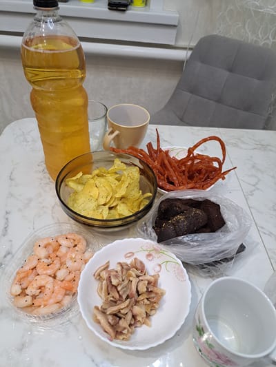 The image shows a table laden with snacks and drinks, likely in a domestic setting in Chernsk, Russia. Dominating the left side is a large, clear plastic bottle filled with a golden-yellow beverage, possibly beer or a similar drink. Beside it is a clear glass with condensation. A beige mug with a handle sits to the right of the glass.
In front of these are various food items. A glass bowl contains a pile of golden potato chips. To its right, a white bowl is filled with thin, reddish, dried meat strips, resembling jerky or a similar snack. Below the potato chips, a clear plastic container holds several cooked shrimp, seasoned with pepper. Next to the shrimp is a white plate with a floral pattern, holding a portion of what appears to be sliced cooked meat, possibly chicken or pork. Behind the plate and to the right, a plastic bag contains dark brown pieces of bread, possibly rye bread, sprinkled with seeds.
In the background, a grey upholstered chair is visible, and part of a white window frame with horizontal blinds is present. The table surface has a marble-like pattern. The overall impression is one of a casual gathering or a personal snack session.