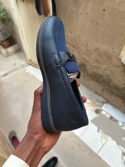 The image displays a close-up shot of a person's hand holding up a single men's loafer shoe. The shoe is predominantly navy blue, crafted from a textured material, likely faux leather, with a contrasting royal blue smooth fabric, possibly suede, on the vamp. A small, metallic silver-toned buckle serves as a decorative element on the front of the shoe. White packing material or tissue paper is visible inside the shoe, indicating it is new or recently prepared for wear.
The loafer features intricate stitching details in a lighter thread, visible along the seams of the upper, around the vamp, and outlining the dark-colored sole which has a thin, lighter grey trim. The hand holding the shoe has dark skin, and a white cuff of a long-sleeved garment is visible at the wrist.
The background is out of focus, suggesting an indoor or semi-outdoor setting, with what appears to be concrete or tiled surfaces. Partial views of other shoes, including a black one and a grey and pink one, are discernible on the floor in the background. The scene is illuminated by natural light, indicating it is daytime. This scene is set in Dan Maliki, Nigeria.