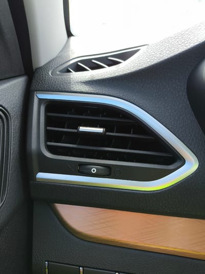 This is a close-up, eye-level, medium shot of the interior of a vehicle, focusing on an air vent. The air vent is black with horizontal slats and a chrome accent. It is framed by a silver and light blue trim that appears to have a yellowish glow on the lower right side. Below the vent is a small black dial with a circular indicator. The textured black dashboard surrounds the vent. To the lower right, a section of faux wood grain trim is visible. The image appears to be taken during the day, with bright light illuminating the scene. There is no indication of the specific location within Tolyatti, Russia, or the presence of any people.