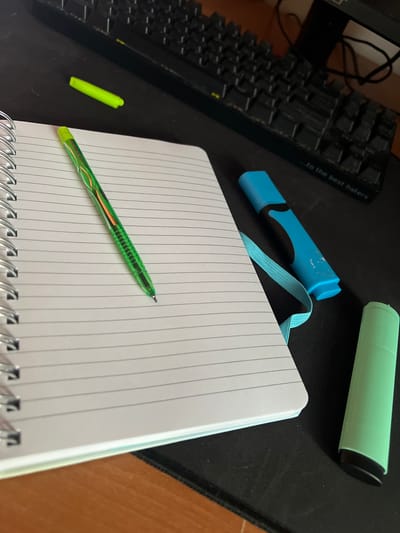 The image depicts a desk with a notebook, pens, and a keyboard. A spiral-bound notebook with lined pages is open on the desk. A green pen with a patterned barrel is resting on the notebook. To the right of the notebook, there is a blue highlighter with a light blue elastic band attached. A green highlighter is also visible on the right side of the desk. In the background, a black keyboard with a message inscribed on it reading "...to the best haters" is partially visible. The desk surface appears to be black, and there is a hint of a wooden surface in the lower part of the image, possibly the edge of the desk. The lighting suggests an indoor setting. There are no people present in the image. The scene is devoid of specific landmarks or context that would indicate its location in Skawina, Poland, or any other city.