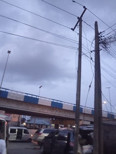 The image depicts an overcast sky over a busy urban street in Port Harcourt, Nigeria. Two prominent wooden utility poles, laden with numerous wires, dominate the foreground. In the mid-ground, a section of an elevated road or bridge, distinguished by white and blue railings, spans across the scene. Below the elevated structure, traffic congestion is evident with several vehicles, including vans and cars, visible on the road. The lighting suggests it is either early morning or late evening. The overall atmosphere is one of an active, densely populated urban environment.