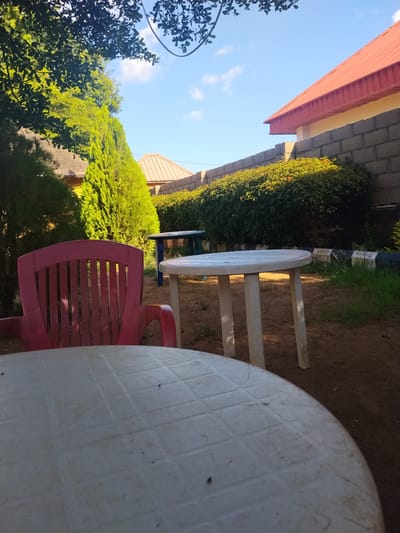 This media file depicts an outdoor seating area, likely a garden or patio, in Mangu Town, Nigeria. The scene is set during the daytime, with bright sunlight casting shadows, suggesting it's either late morning or early afternoon. The weather appears clear and pleasant, with a blue sky dotted with a few wispy clouds.
Dominating the foreground is a large, white plastic table, its textured surface filling much of the frame. Behind it, a red plastic chair is positioned, its slatted back visible. Further back, another white plastic table with three legs is visible, along with a smaller, blue table in the background. The ground is covered in reddish-brown soil, with patches of dry grass.
The backdrop features lush green foliage, including tall, conical trees and a thick, manicured hedge. Beyond the hedge, a low brick wall separates the seating area from a building with a red, corrugated metal roof and a pale yellow exterior wall. The architectural style and the dense vegetation are indicative of a tropical or subtropical climate, consistent with the specified location in Nigeria.
There is no discernible activity or event taking place; the scene appears to be a still shot of an empty outdoor space, possibly set up for casual use. The overall mood is one of quiet repose, with the bright sunlight and greenery creating a cheerful and inviting atmosphere. No text is visible in the image.
