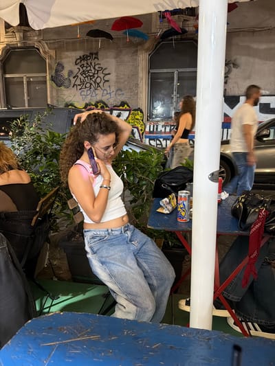 The image captures an outdoor evening scene on a street in Catania, Italy. A young woman with long, curly brown hair, wearing a white cropped tank top with pink trim and light blue jeans, is seated at a blue rectangular table, holding a purple-cased smartphone to her left ear with her left hand while her right hand is raised to her hair. She wears glasses and multiple bracelets. To her left, another woman is partially visible from the back, seated at an adjacent table.
On the blue table, a red Bull Dog energy drink can, a glass of amber liquid, and another red beverage can are visible, alongside a black shoulder bag. A prominent white pole stands centrally on the right, partially obscuring the background. In the mid-ground, a blurred woman in a black crop top and light jeans walks left-to-right, while a blurred man in a white t-shirt and blue jeans walks right-to-left behind the white pole, near a silver car.
The background features a multi-story building wall heavily covered in black graffiti, including "CAROTA BANG ROSA LATTOC ZONA SPA PIG" and "VIVA L'ATANAZ", along with other tags and abstract designs. A window with a metal grate is visible on this wall. Numerous small, inverted umbrellas in red, orange, black, blue, and pink are suspended overhead as decoration. Potted green plants are arranged around the seating area. The ground under the tables is a mix of dark pavement and a green surface. In the bottom right, the legs and feet of a person wearing dark jeans and white Adidas sneakers are visible, with a red bandana tied around one leg.