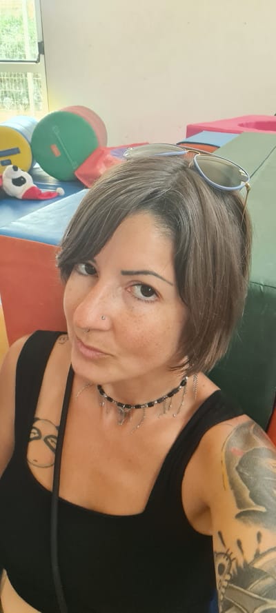 A woman with short grey hair, freckles, and a nose piercing is pictured in a selfie. She wears sunglasses atop her head and a black tank top. A silver and black necklace adorns her neck. A tattoo is visible on her left upper arm. In the background, there are colorful soft play objects, and a stuffed animal wearing a Santa hat. The environment appears to be indoors, likely in the city of Lachish Regional Council, Israel.