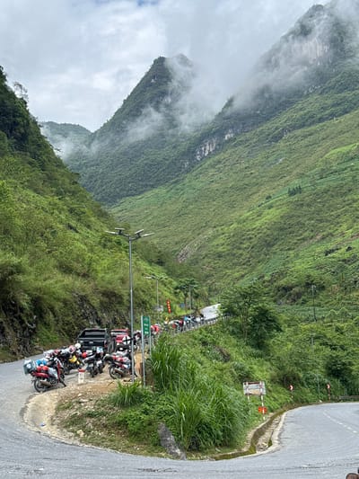 The image shows a roadside scene in the Yên Minh Commune, Vietnam, with steep, green, mountainous terrain in the background partially obscured by low-lying clouds. A narrow asphalt road curves to the right of the frame. Parked along the roadside is a dense cluster of motorcycles and at least one black pickup truck. Utility poles and signage are visible along the road. The surrounding landscape is covered in lush vegetation.