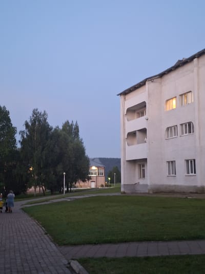The image depicts an outdoor scene in Атамановка, Russia, at dusk. In the foreground, a paved walkway leads away from the viewer to the left. A woman in a blue dress is pushing a stroller with a child, and another child is walking beside them. To the right of the walkway is a well-maintained lawn.
Dominating the right side of the frame is a three-story building with a white facade. The building has balconies on the upper two floors and several windows. One window on the top floor emits a warm yellow light, indicating an interior light source. The roofline of this building is visible at the top.
In the background, to the left of the main building, is another, smaller building with a light-colored facade and a darker roof. This building also has windows, and one appears to be illuminated with a yellow light. A lamppost stands on the lawn in front of this building. Behind the buildings, a dense line of trees, including birch trees, creates a dark green backdrop against the pale blue evening sky. The distant landscape shows rolling hills. The overall atmosphere is peaceful and quiet, characteristic of a residential area in the early evening.