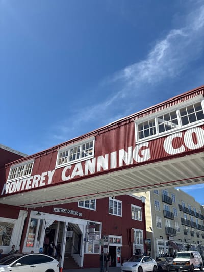 The image captures an outdoor daytime scene on Cannery Row in Monterey, California, United States. The central focus is a two-story building complex constructed primarily of dark red corrugated metal with white trim and numerous white-framed windows. An overhead pedestrian bridge, also featuring red corrugated metal, spans across the scene with "MONTEREY CANNING CO" written in large white letters. The ground floor of the main building features multiple storefronts with white wooden facades, one prominently marked "MONTEREY CANNING CO" and "711". Several individuals are visible on the sidewalk and entering the building. Parked on the street in front of the building are a white Tesla Model X (partially visible on the left), a white Tesla Model Y, a dark grey SUV, and a white van. To the right of the red building, a taller, multi-story beige building with multiple balconies and windows is visible. The sky is clear and blue with sparse, wispy white clouds.