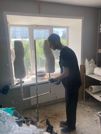 The image shows a person in a workshop, likely involved in sculpting or mold-making. The subject, wearing a dark t-shirt and dark pants with blue Crocs, is leaning over a plaster cast. The cast is shaped like a torso and is mounted on a metal stand with rough twine binding it. Another similar cast is positioned above it, also on a stand.

The workshop appears cluttered with materials. Bags of white powder are stacked in the background, along with various tools and what looks like construction debris on the floor. A large window allows natural light into the room, suggesting it's daytime. Through the window, a residential building and trees are visible, hinting at an urban or suburban setting. The presence of the plaster casts and the materials implies a creative or manufacturing process is underway. The overall impression is of a functional, perhaps somewhat messy, workspace. There is no visible text in the image.
