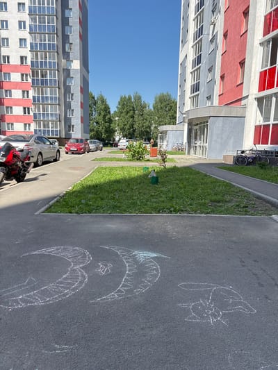 The image depicts a courtyard area in front of apartment buildings in Kemerovo, Russia. Two multi-story apartment buildings, one primarily red and gray, the other gray and red, are visible. A patch of green grass is situated between a paved walkway and the foreground asphalt. On the asphalt, drawings made with chalk are visible, including what appear to be crescent moons and a cat-like creature. Parked cars and a motorcycle are present in the background. Several trees line the area behind the buildings. The sky is clear and blue. There are no people actively engaged in any activity in the foreground; however, a few individuals are faintly visible in the background near the building entrances. The scene appears to be a residential area on a sunny day.