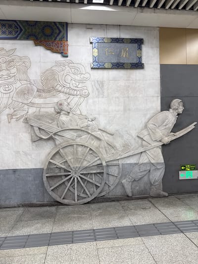 The image depicts a large bas-relief carving on a marble wall, located in Dongcheng District, China. The carving features a historical scene with two main elements. On the left, two stylized, dragon-like creatures are depicted, possibly in motion, with flowing manes. To the right of these creatures, a woman is seated in a two-wheeled cart. She is dressed in traditional attire and holds a large, round object. The cart is being pulled by a man, also dressed in traditional clothing, who is shown in profile, leaning forward as he pushes the cart's handles. The carving is rendered in a monochromatic, stone-like material against the white marble background. Above the carving, a rectangular plaque with Chinese characters and decorative borders is affixed to the wall. The environment appears to be an indoor public space, possibly a subway station or an underpass, with a grey, textured wall section to the right of the carving. Green exit signs are visible on this grey wall section. The floor in front of the carving is tiled with a speckled pattern, and a tactile paving strip runs parallel to the wall.