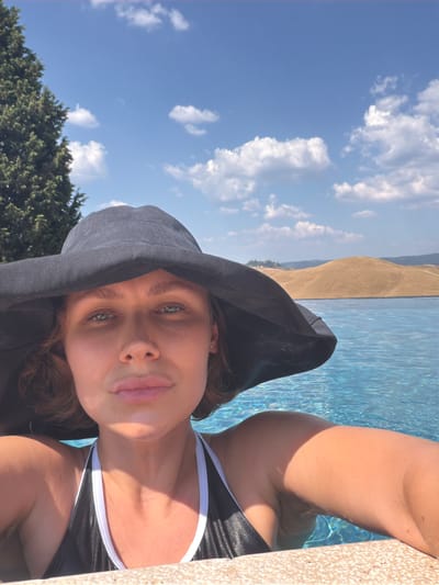 A woman is taking a selfie by the pool with a large black hat. The woman is wearing a black and white swimsuit. The water is blue and clear, and there are rolling hills in the background. The sky is blue with white clouds. The scene is likely outdoors, possibly in Asciano, Italy, given the hilly landscape and clear sky which are characteristic of the Tuscan region.